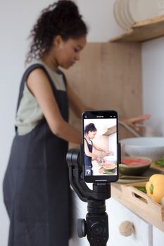 Side view of a woman filming a cooking video with a smartphone in the kitchen.