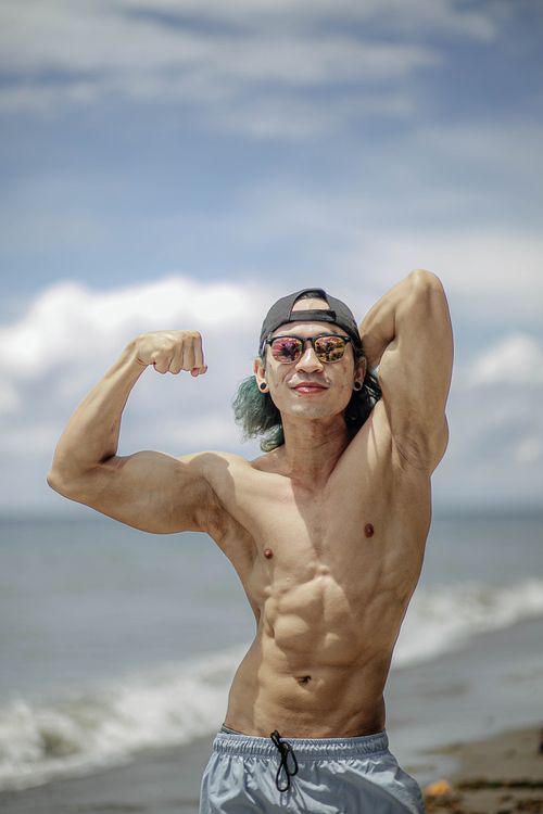 Free Shirtless muscular man flexing on a sunny beach day, wearing sunglasses and cap. Stock Photo