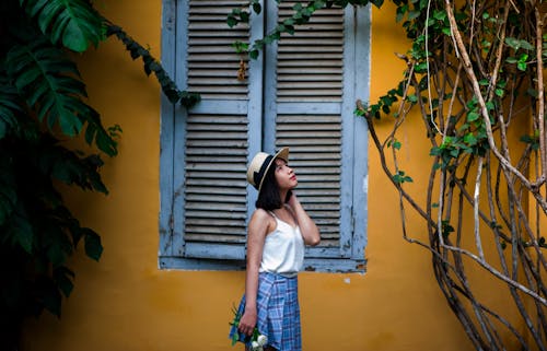 Photo of Woman Wearing White Sleeveless Top and Hat Holding Flowers Near Window