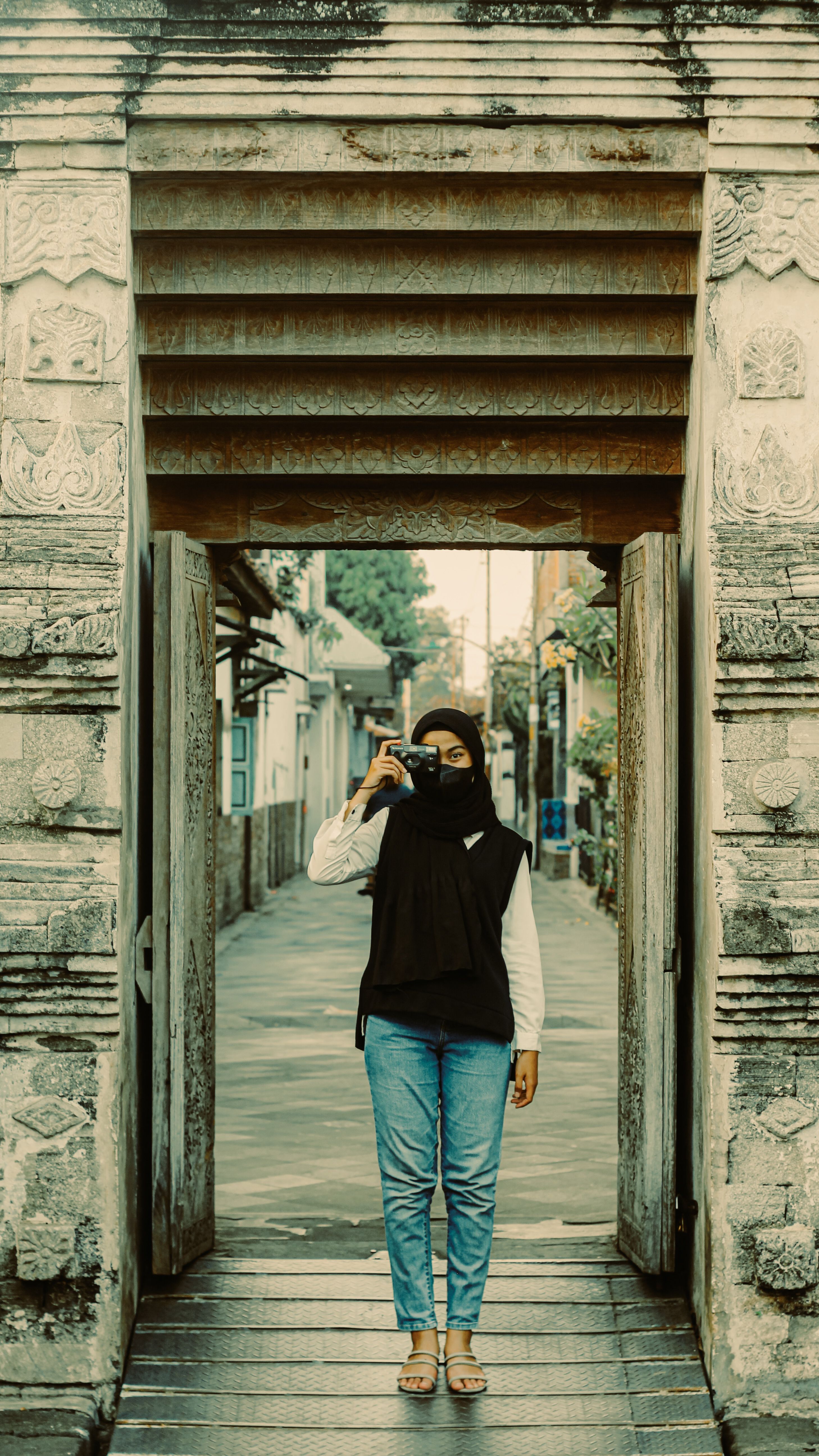 A woman wearing a hijab is taking a photo with a vintage camera under a stone arch outdoors.