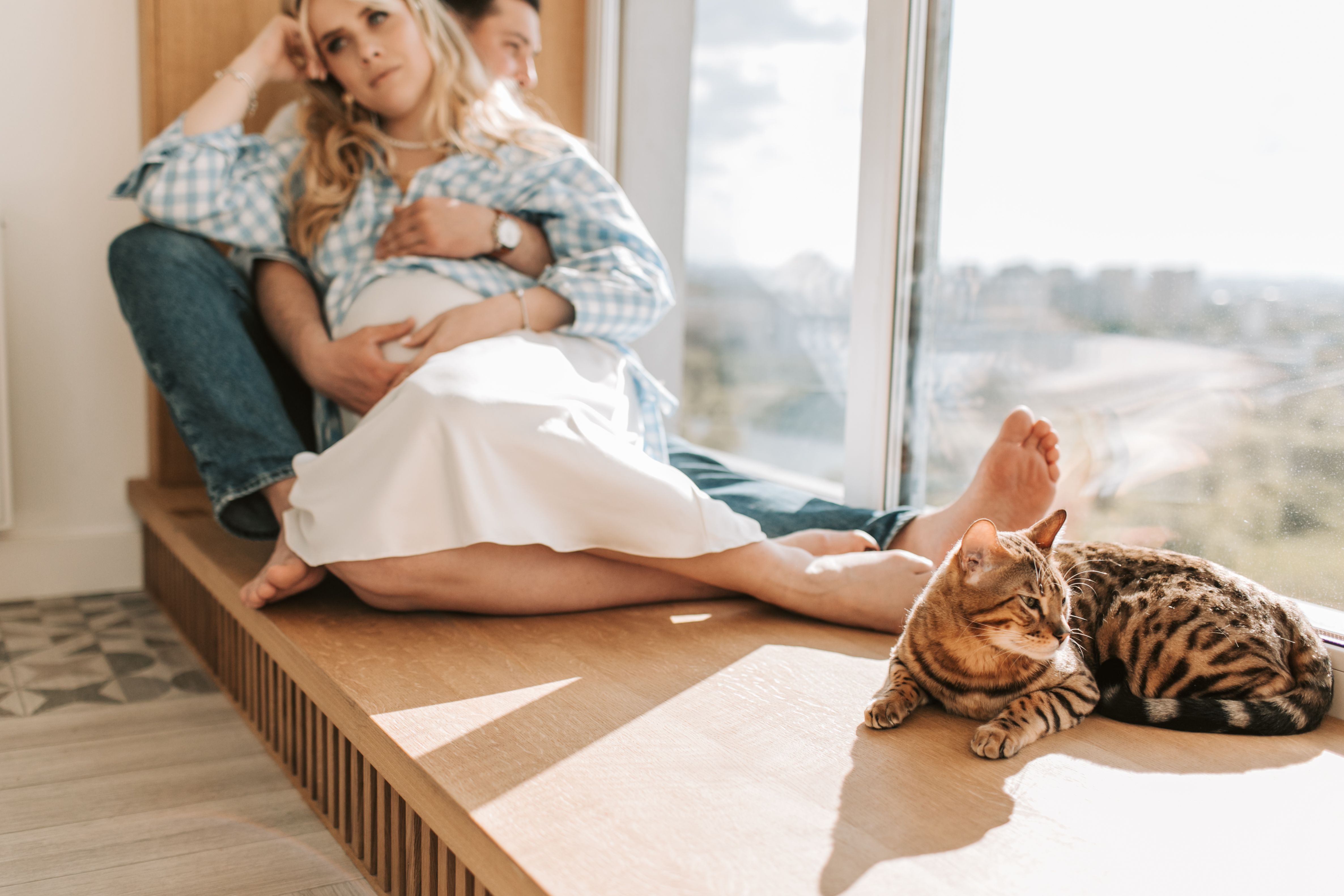 Pregnant couple embraces while sitting indoors with their cat by a window.