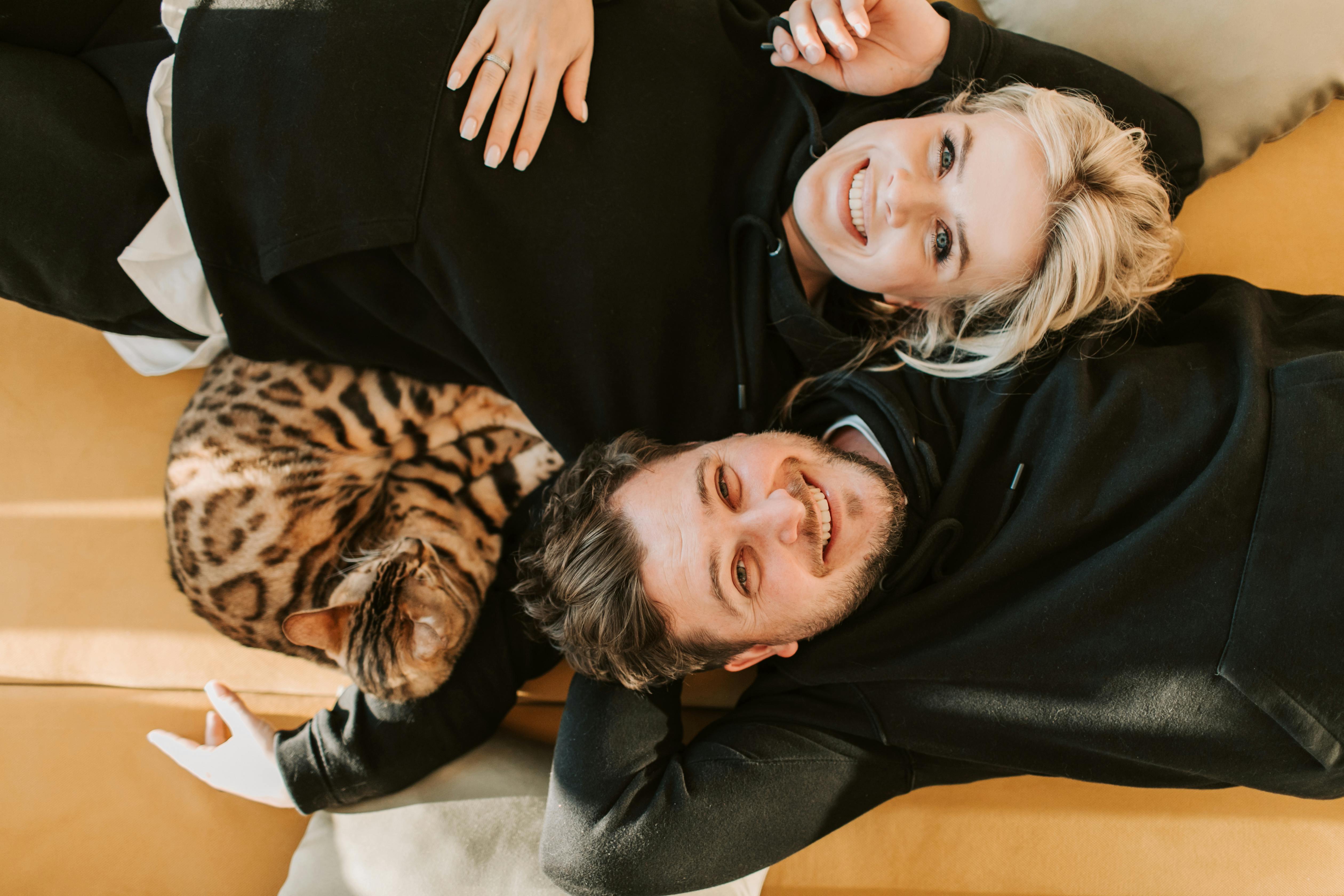 Smiling couple relaxing with Bengal cat indoors, embodying love and comfort.