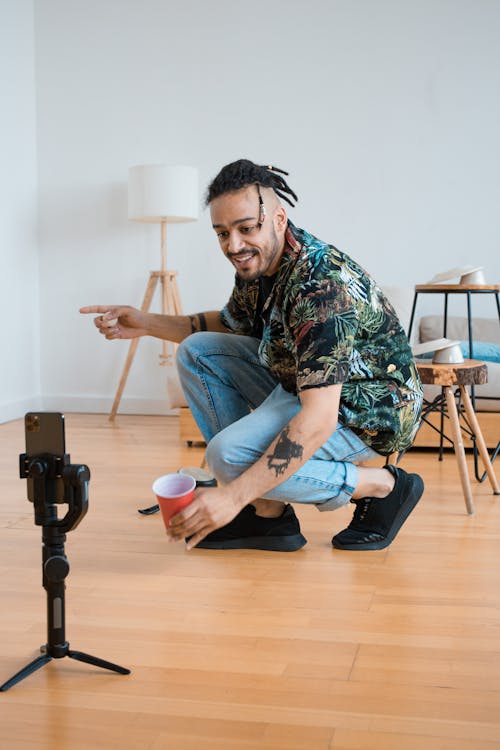 Man in a printed shirt and jeans holding a red cup in front of a phone.