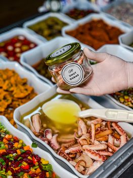 Close-up of a glass jar with pickled octopus held above a selection of gourmet foods.