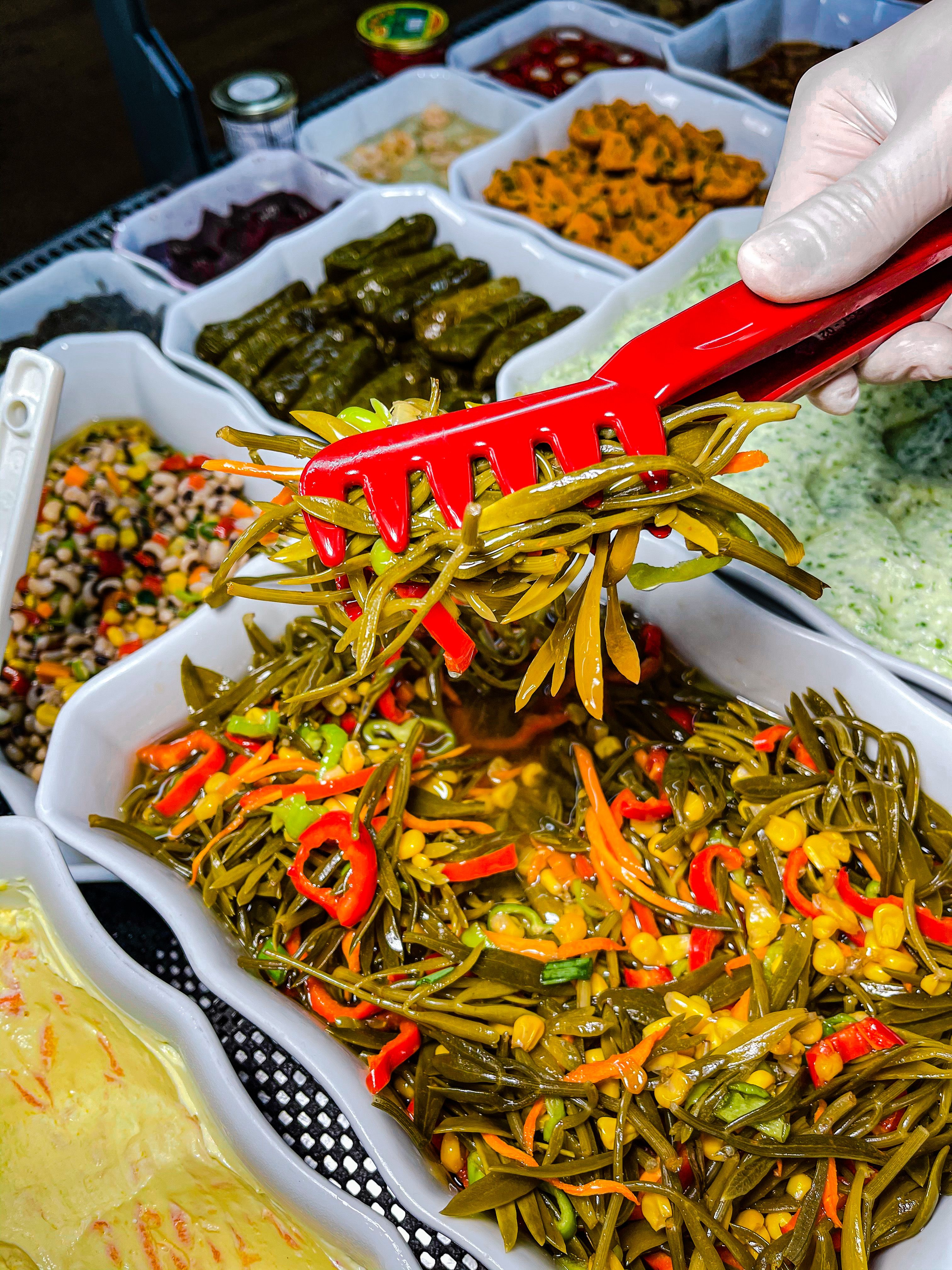 Close-up of a hand with red tongs picking colorful vegetable salad in deli display.