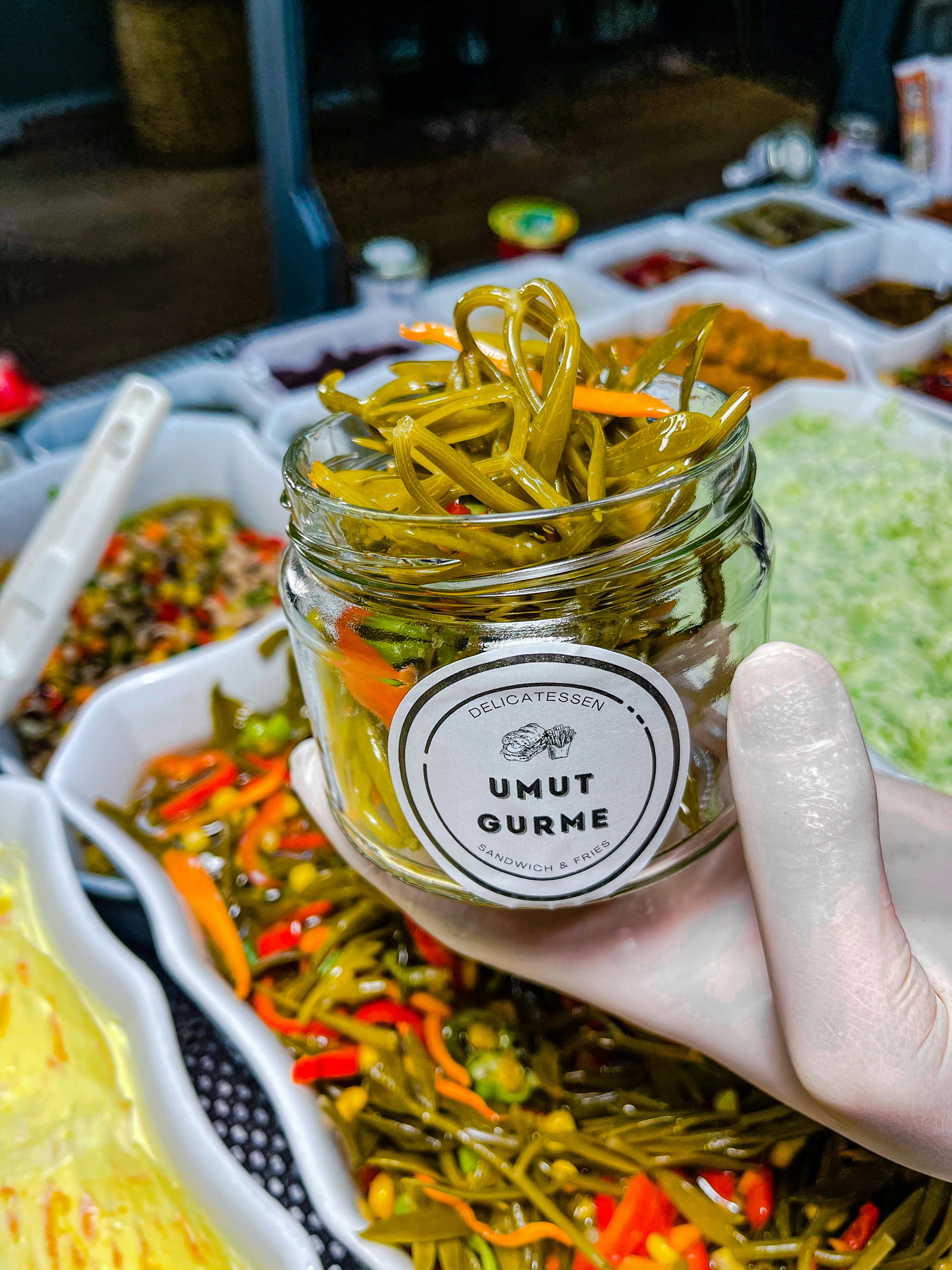 Gloved hand holding a jar of gourmet pickled peppers in a vibrant food display setting.