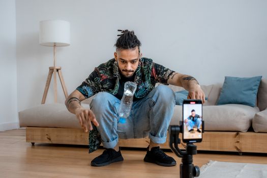 A young man focuses on a bottle flip challenge while filming content at home, capturing it on a smartphone.