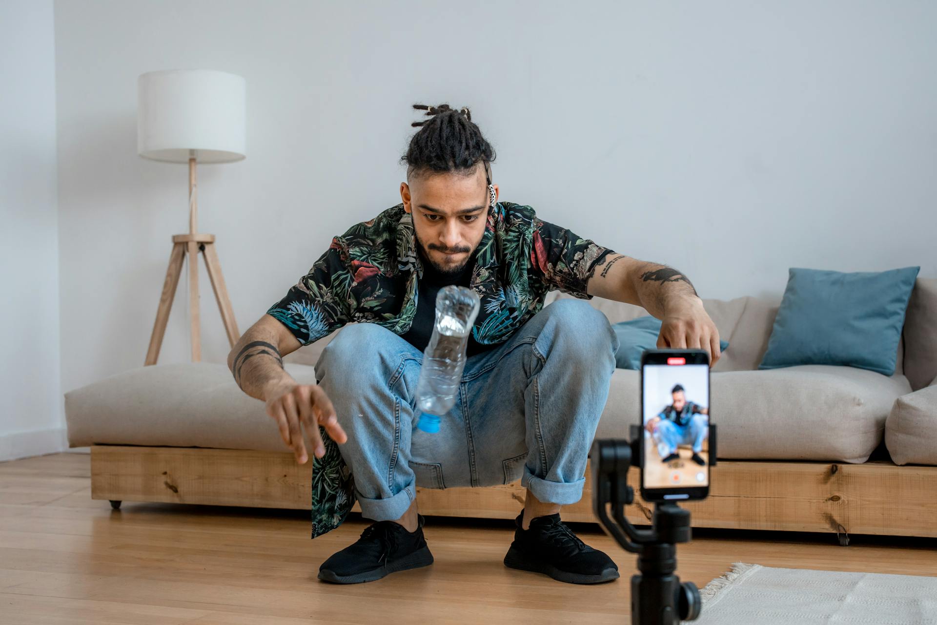 A young man focuses on a bottle flip challenge while filming content at home, capturing it on a smartphone.