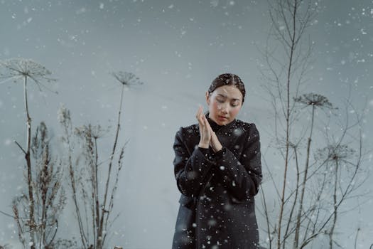 A serene woman in a black coat stands amidst falling snow with dried twigs, embracing winter's beauty.
