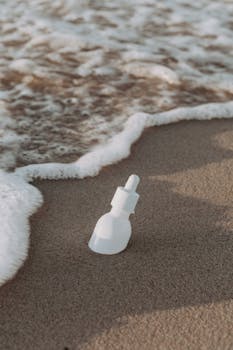 White cosmetic dropper bottle on sandy shore, touched by gentle waves.