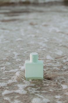 Minimalist perfume bottle resting on the sandy beach surrounded by ocean waves.
