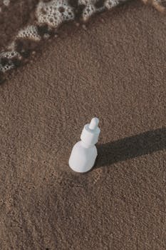 White cosmetic bottle in sand with ocean waves, offering natural beauty care theme.