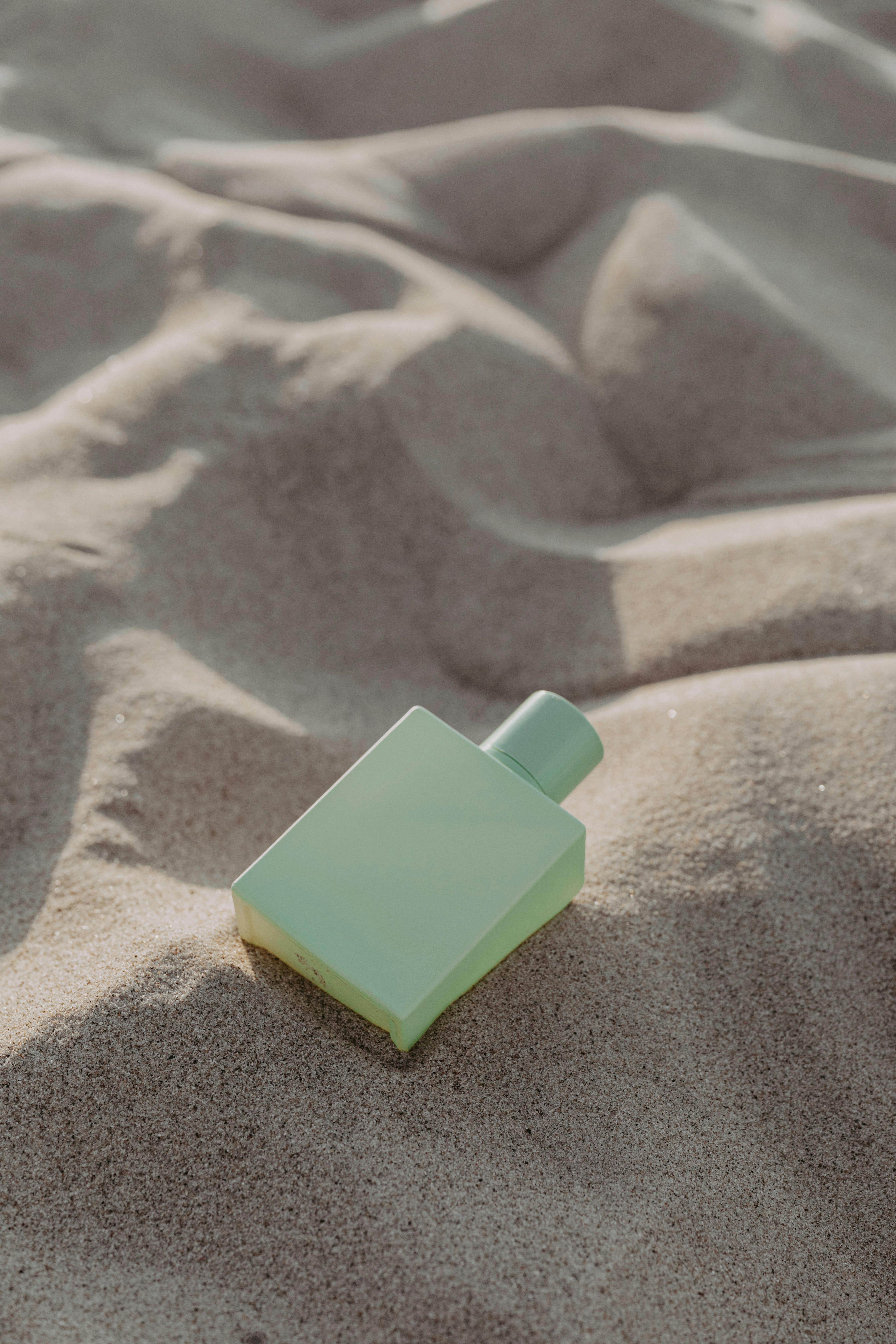 A pastel green perfume bottle rests elegantly on rippled sand under natural lighting.