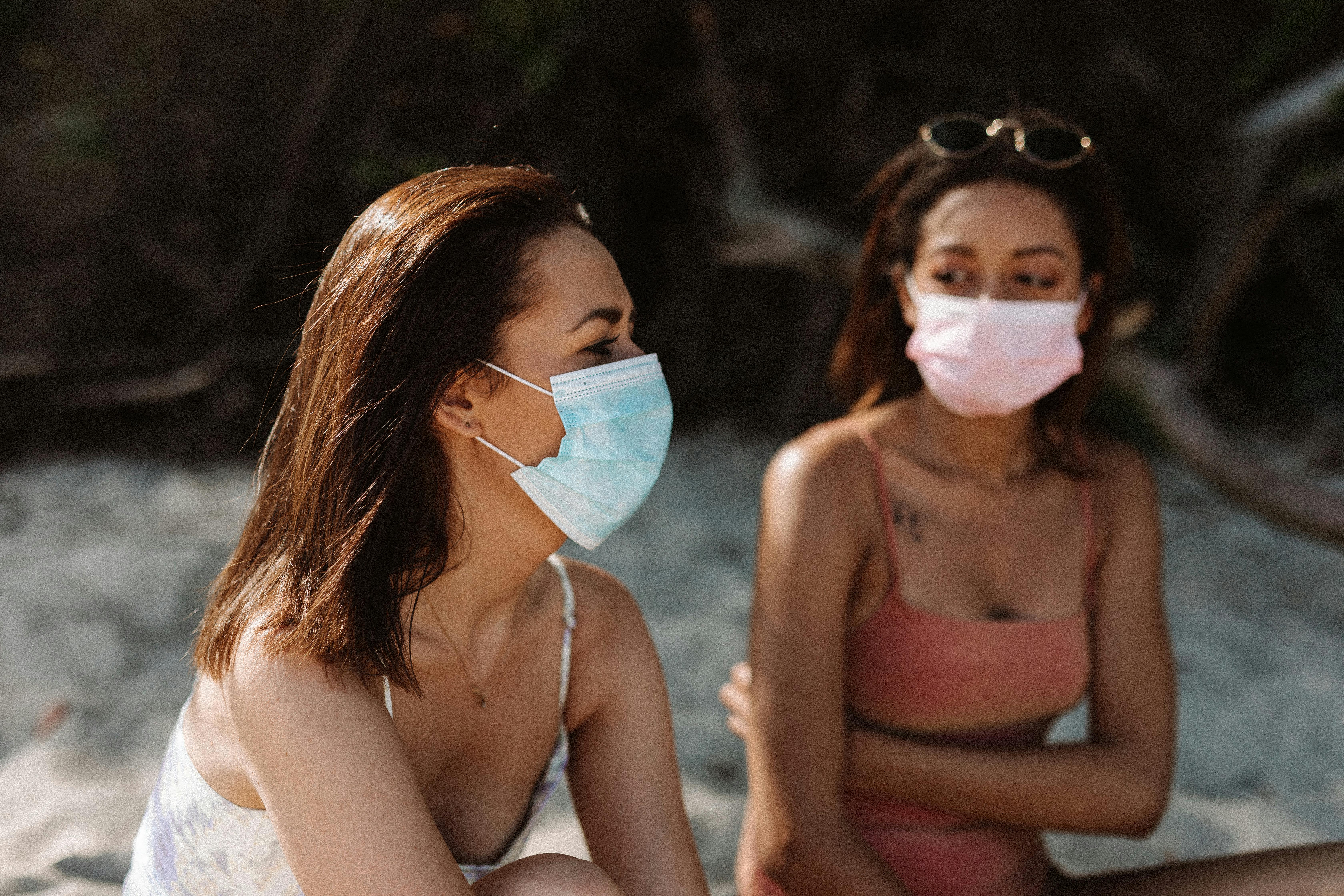 Two women wearing face masks relaxing at a sunlit beach, embracing the new normal.