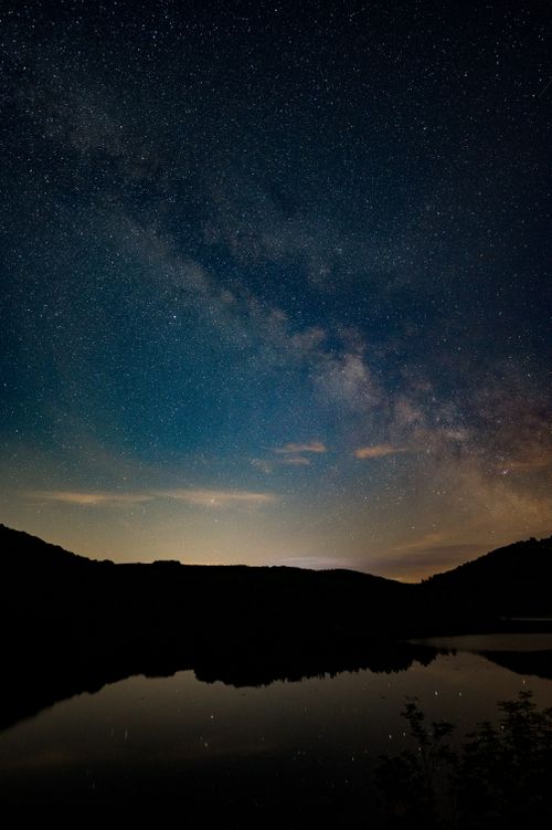 Free A breathtaking starry night sky reflecting over a tranquil lake surrounded by silhouettes of hills. Stock Photo