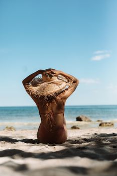 Back view of a woman sunbathing on a sandy beach with ocean waves and clear blue sky.