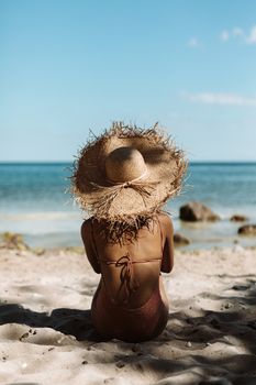 Woman sitting on the beach in a swimsuit and straw hat, enjoying summer relaxation by the ocean.