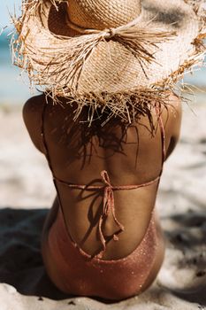 Woman in swimsuit with straw hat sitting on sandy beach enjoying vacation.