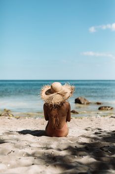 A peaceful scene of a woman sitting on the beach wearing a straw hat, with a clear blue sky and ocean view.