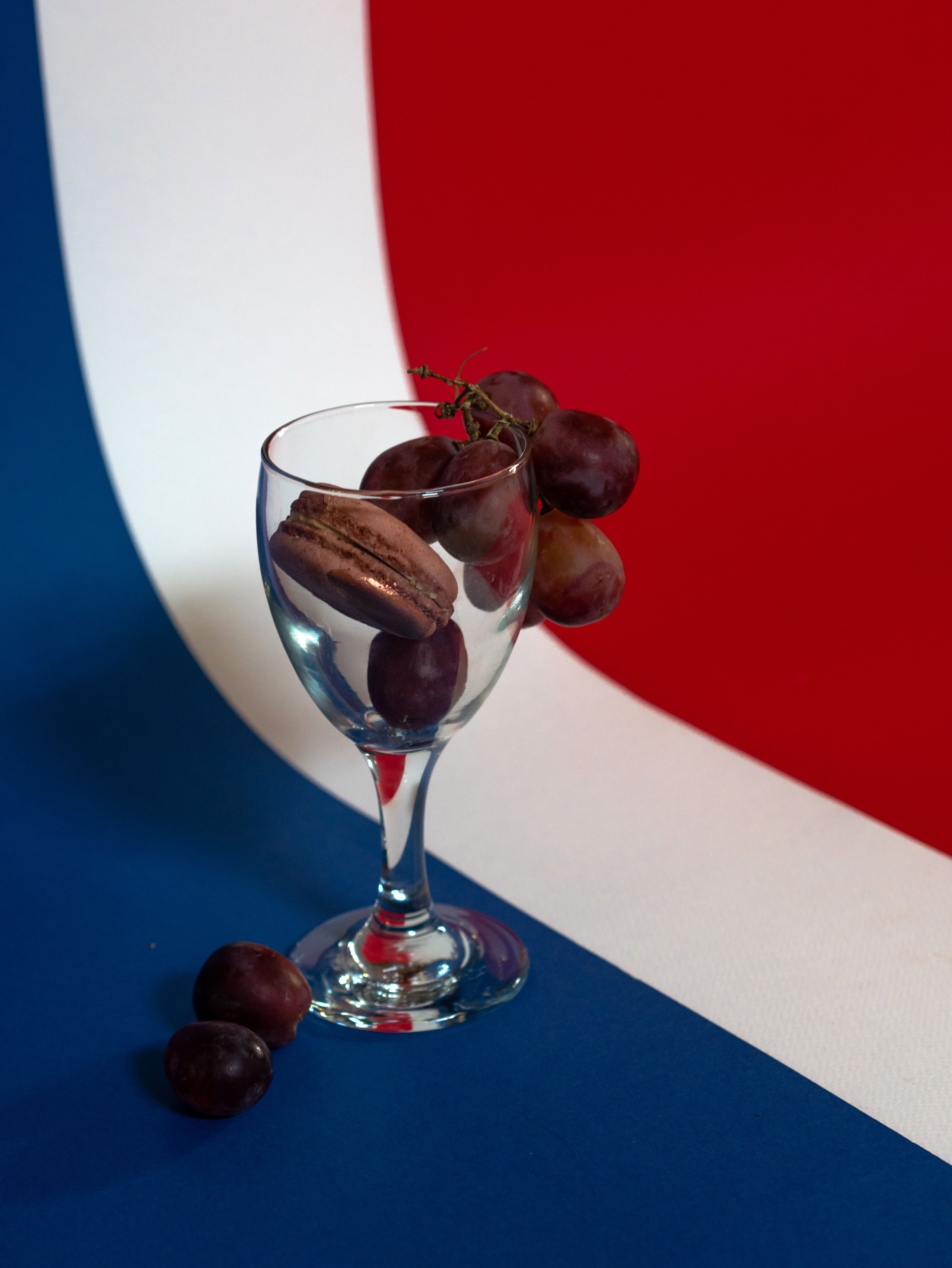 Elegant still life of a wine glass with red grapes and a chocolate macaron on a French-themed backdrop.