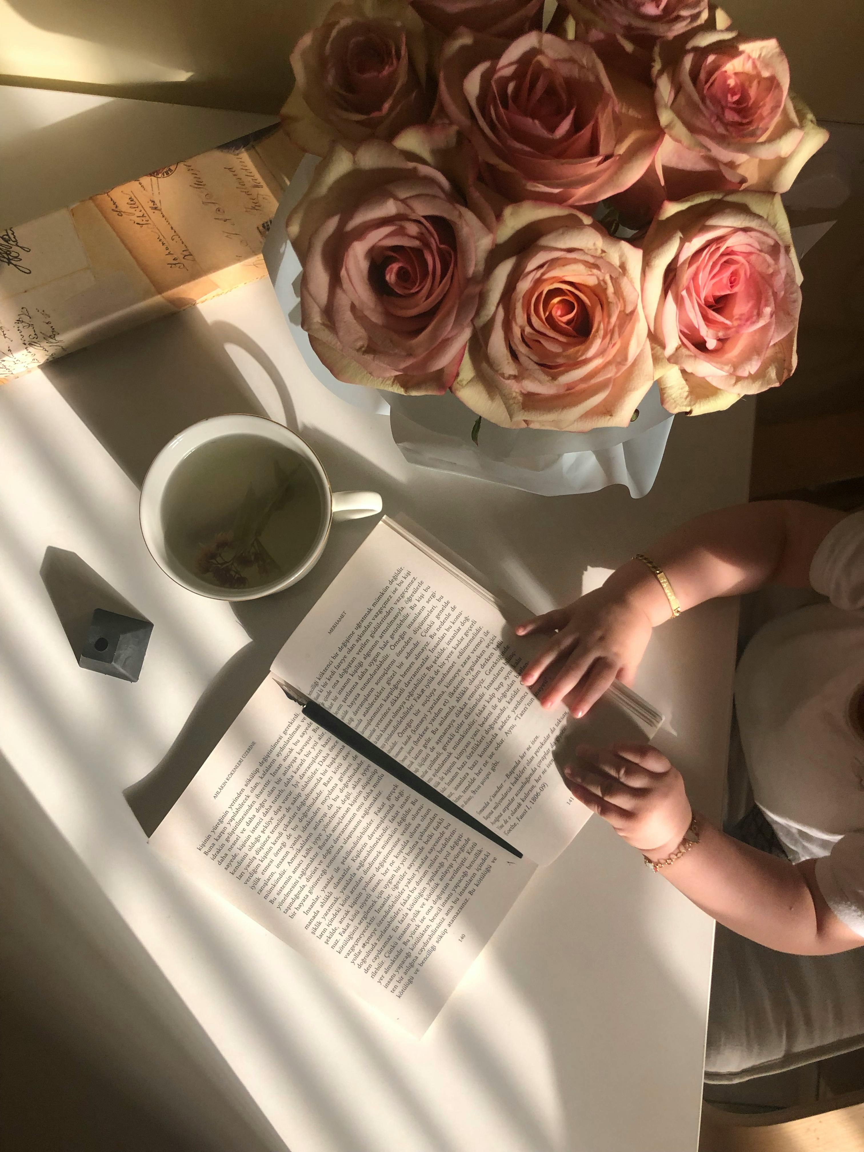 A baby touches a book on a table with roses and tea in a warmly lit room.