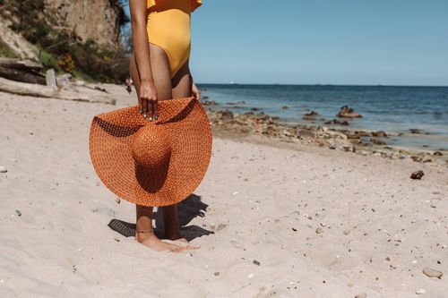 Free A woman stands on a sandy beach holding a large straw hat under the bright summer sun. Stock Photo