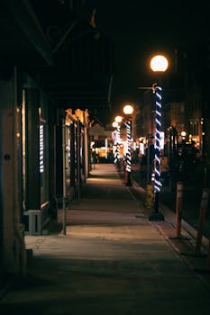A dimly lit city sidewalk at night with decorative lighting and street lamps, creating a moody atmosphere.