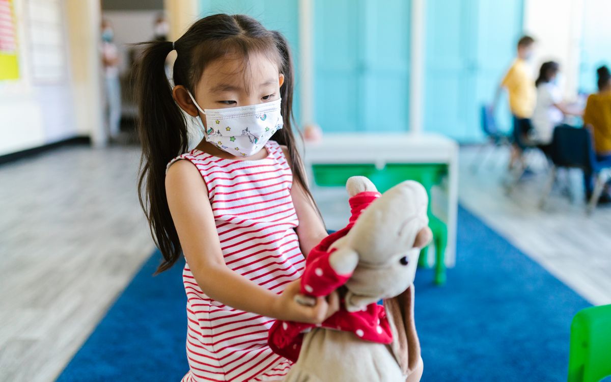 A young girl with pigtails wearing a face mask holding a stuffed toy in a classroom setting.