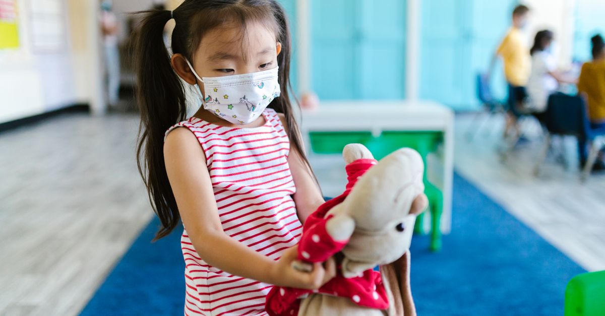 Photo by RDNE Stock project A young girl with pigtails wearing a face mask holding a stuffed toy in a classroom setting.