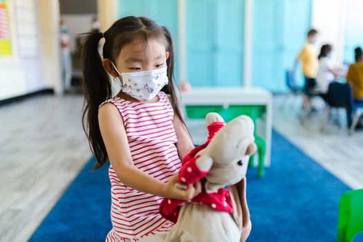 Photo by RDNE Stock project A young girl with pigtails wearing a face mask holding a stuffed toy in a classroom setting.