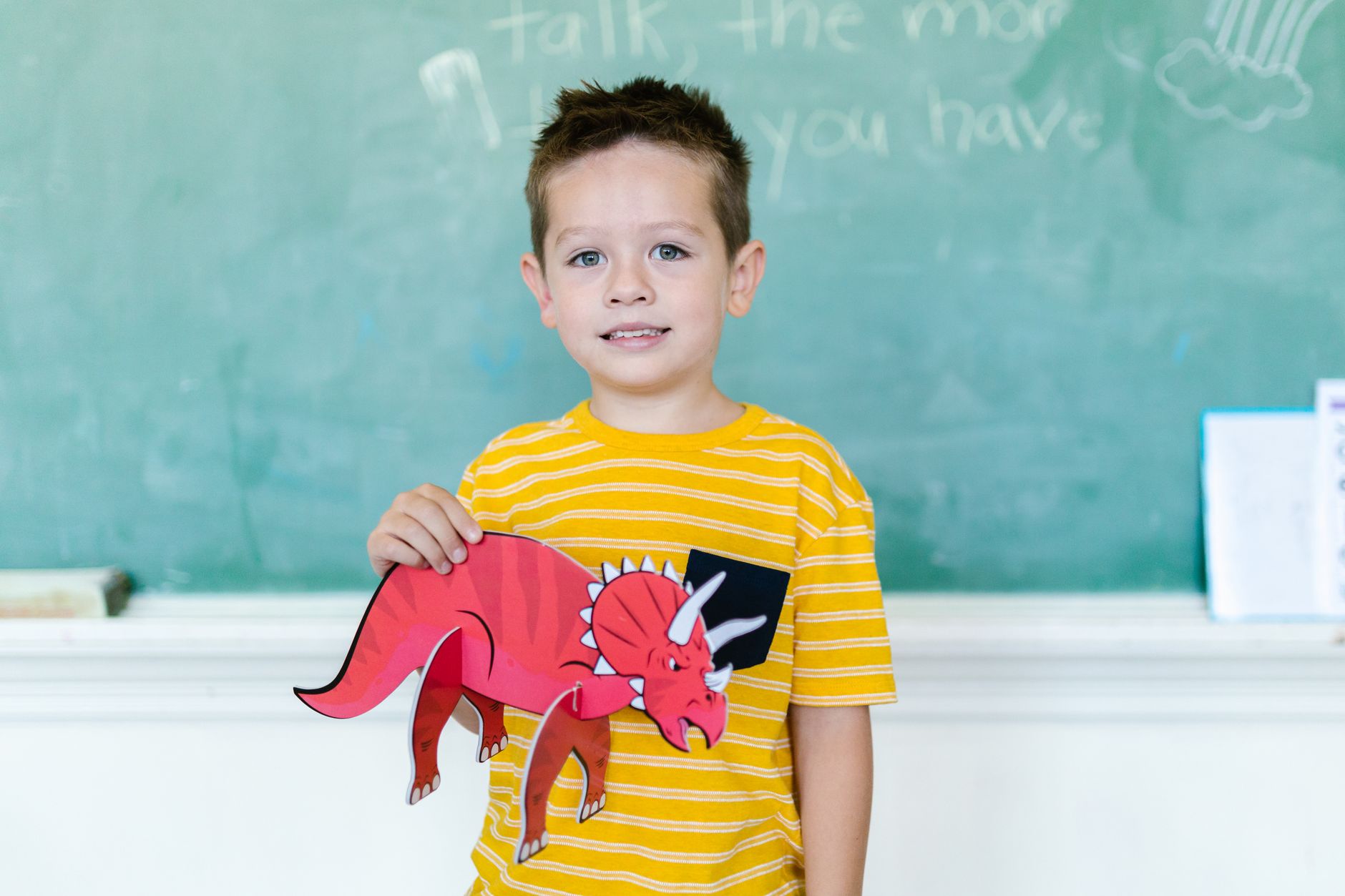 A young boy proudly displays a red triceratops paper cutout in a classroom setting.