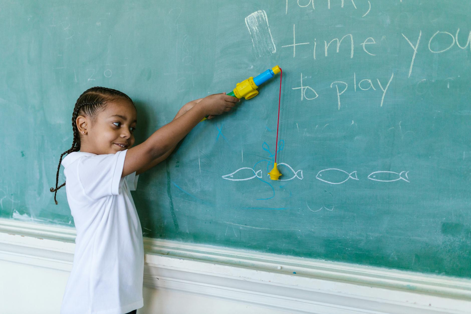 A young boy with braided hair playing with a toy fishing rod by a blackboard in a classroom setting.