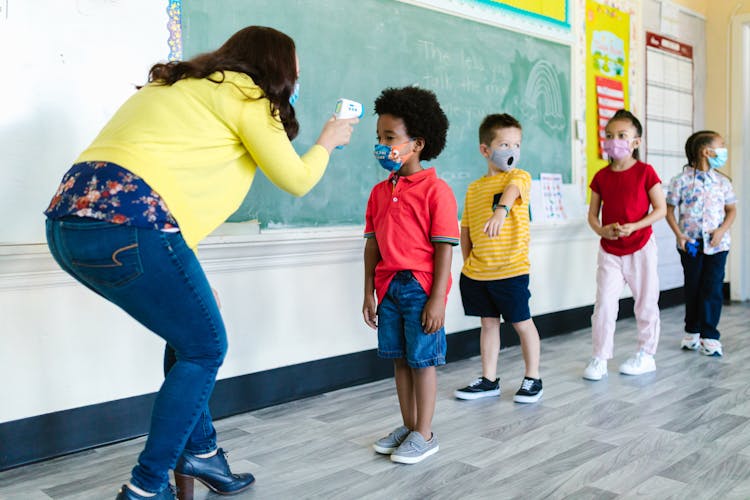 Teacher In Yellow Cardigan Measuring Temperature Of A Boy In Face Mask In The Classroom