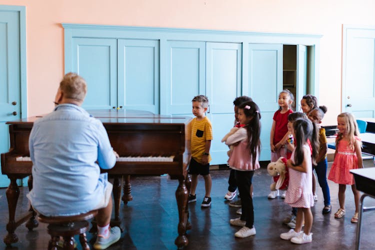 A Group Of Kids Standing Near The Man Playing Piano 