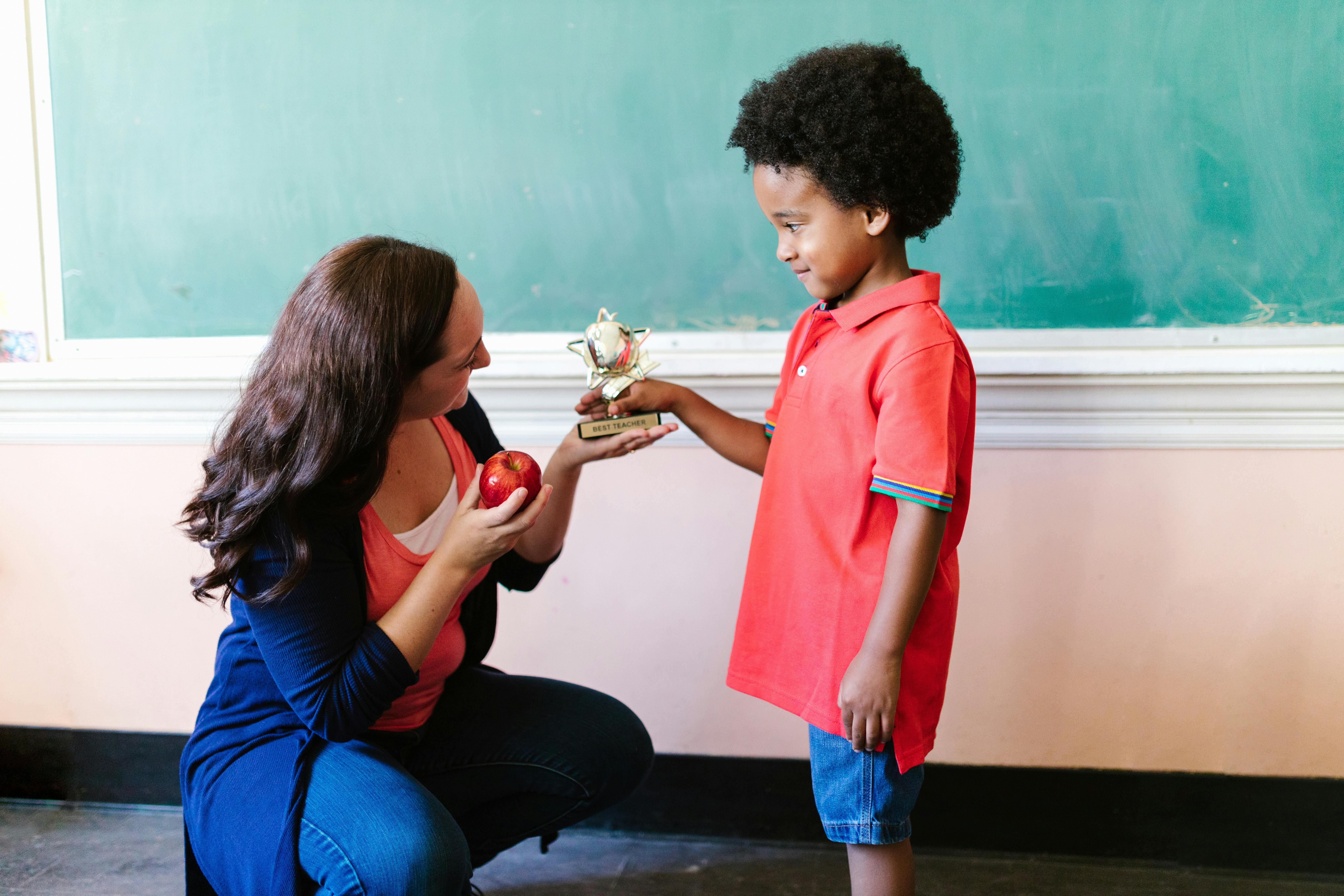 A Kid handing Out a Best Teacher Trophy to a Woman Holding Apple