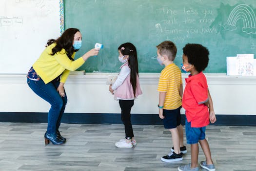 A teacher in a mask checks students' temperatures with a non-contact thermometer in a classroom setting.