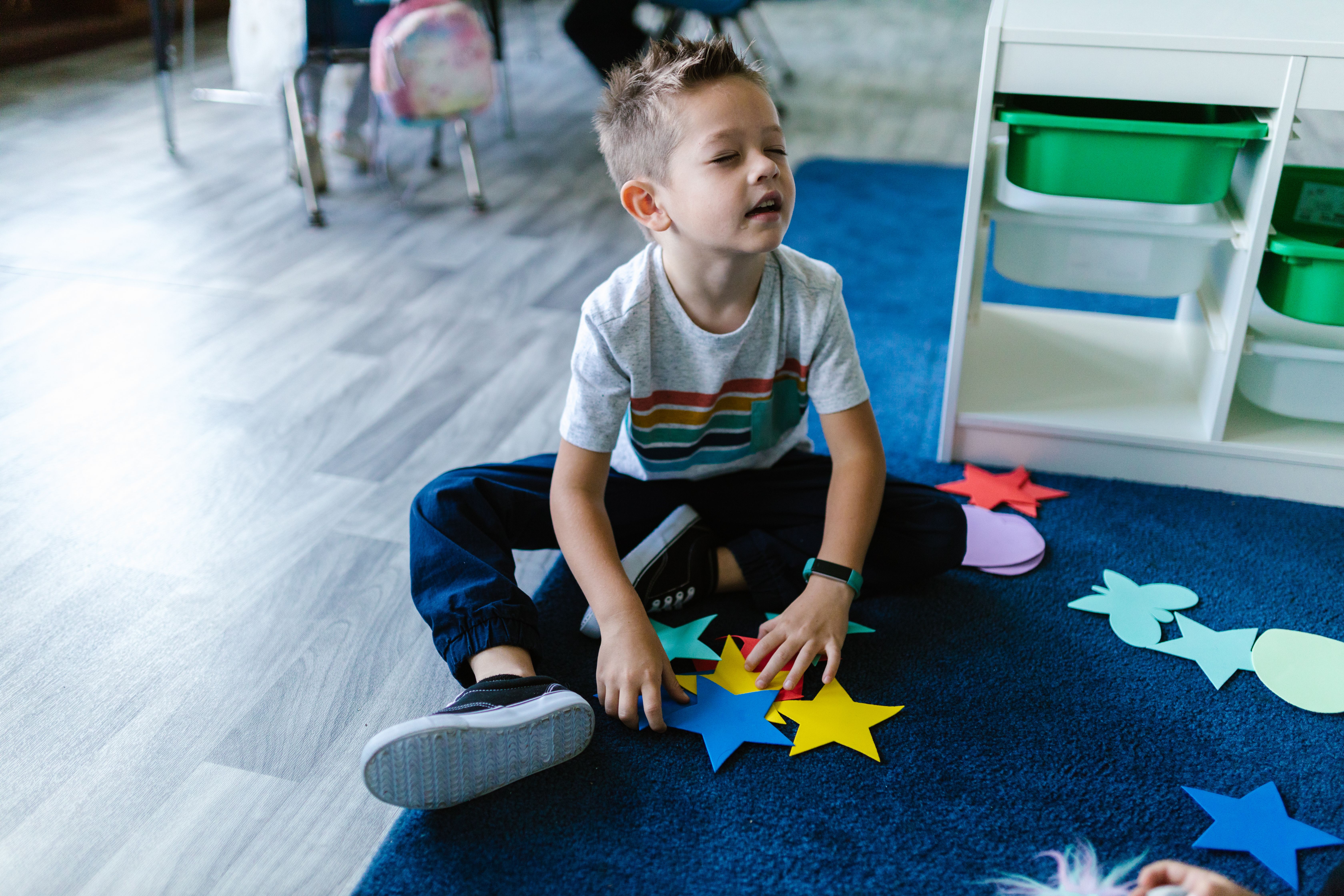 Un jeune garçon est assis par terre, les yeux fermés, jouant avec des formes en papier colorées dans une salle de classe.