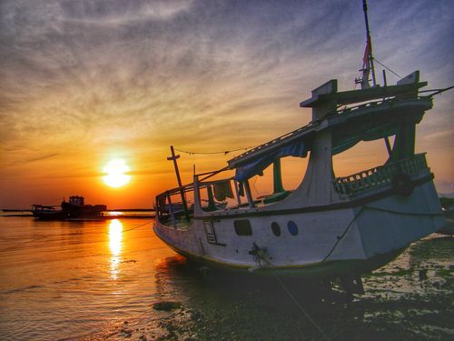Free A scenic view of a rustic boat on the seashore at sunset, casting warm reflections on the ocean. Stock Photo