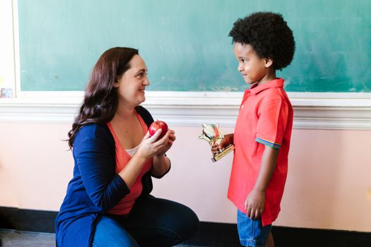 A teacher presents a student with an apple and trophy in a classroom setting.