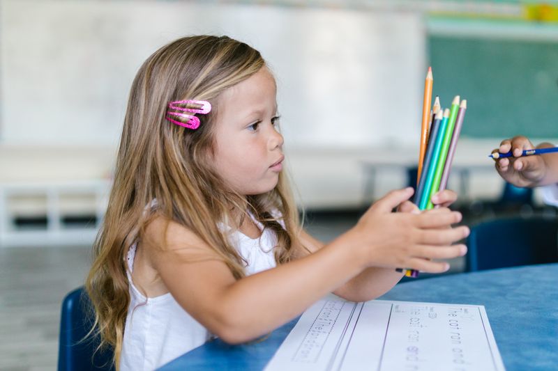 A young girl holds colored pencils while sitting at a desk in a classroom setting.