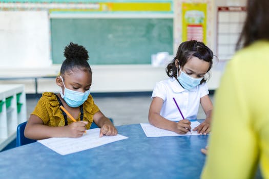 Young children in masks write in a classroom, focusing on schoolwork during the pandemic.
