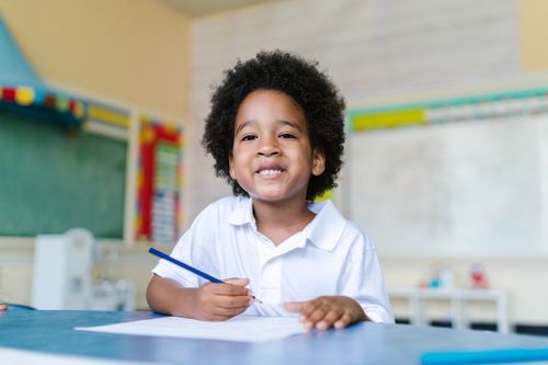 Free Happy child with afro hair sitting in a classroom, holding a pencil and smiling. Stock Photo