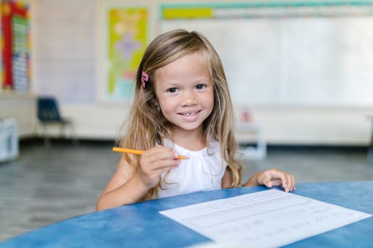 A young girl smiles while working on schoolwork in a bright classroom.
