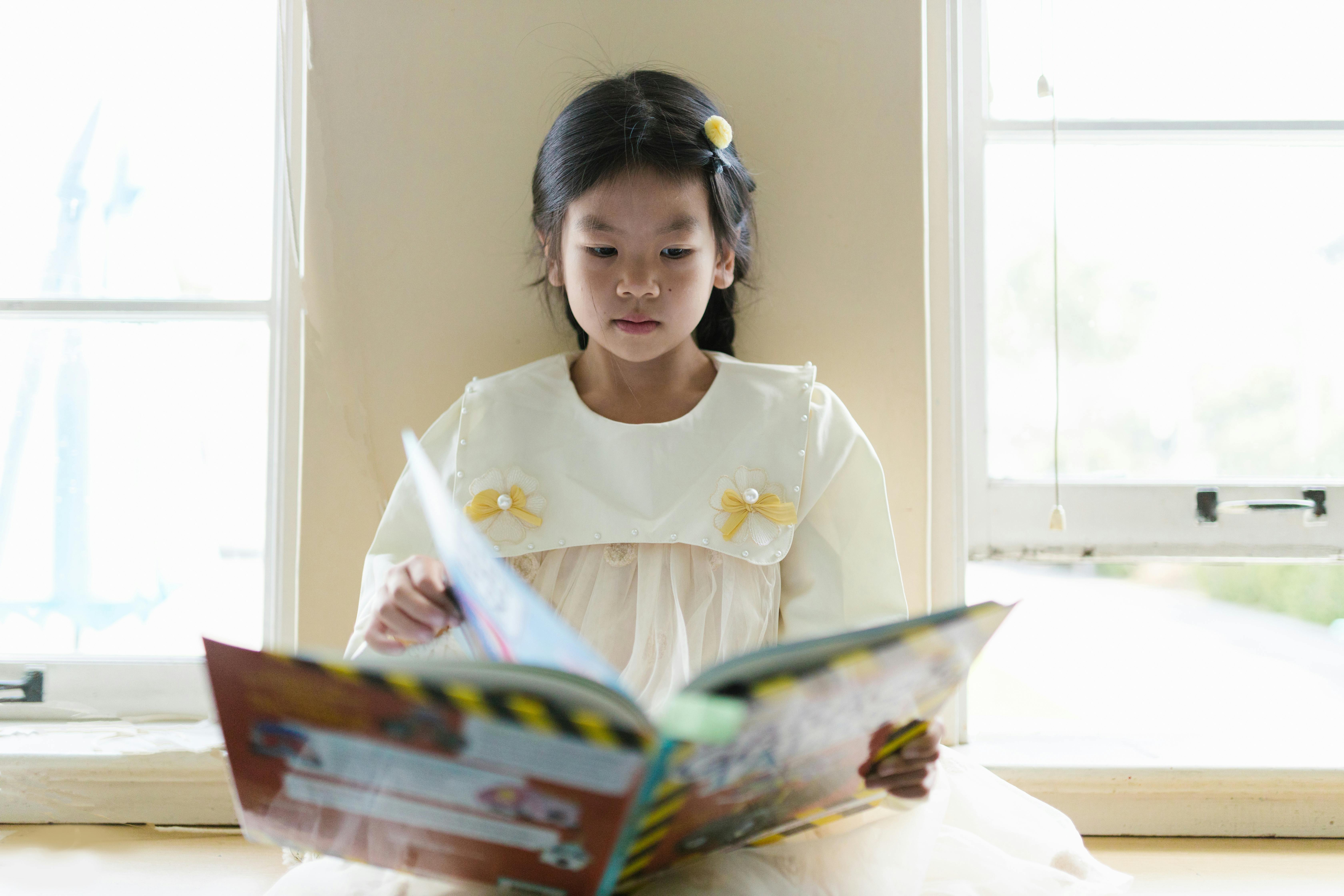 Free A young girl in a dress enjoys reading a book by a bright window, capturing a moment of serene concentration indoors. Stock Photo