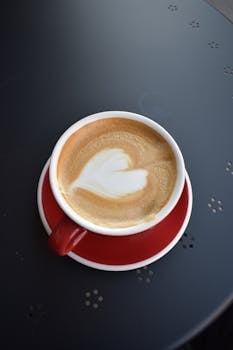 Top view of a creamy latte with heart art in a red cup and saucer on a dark table.