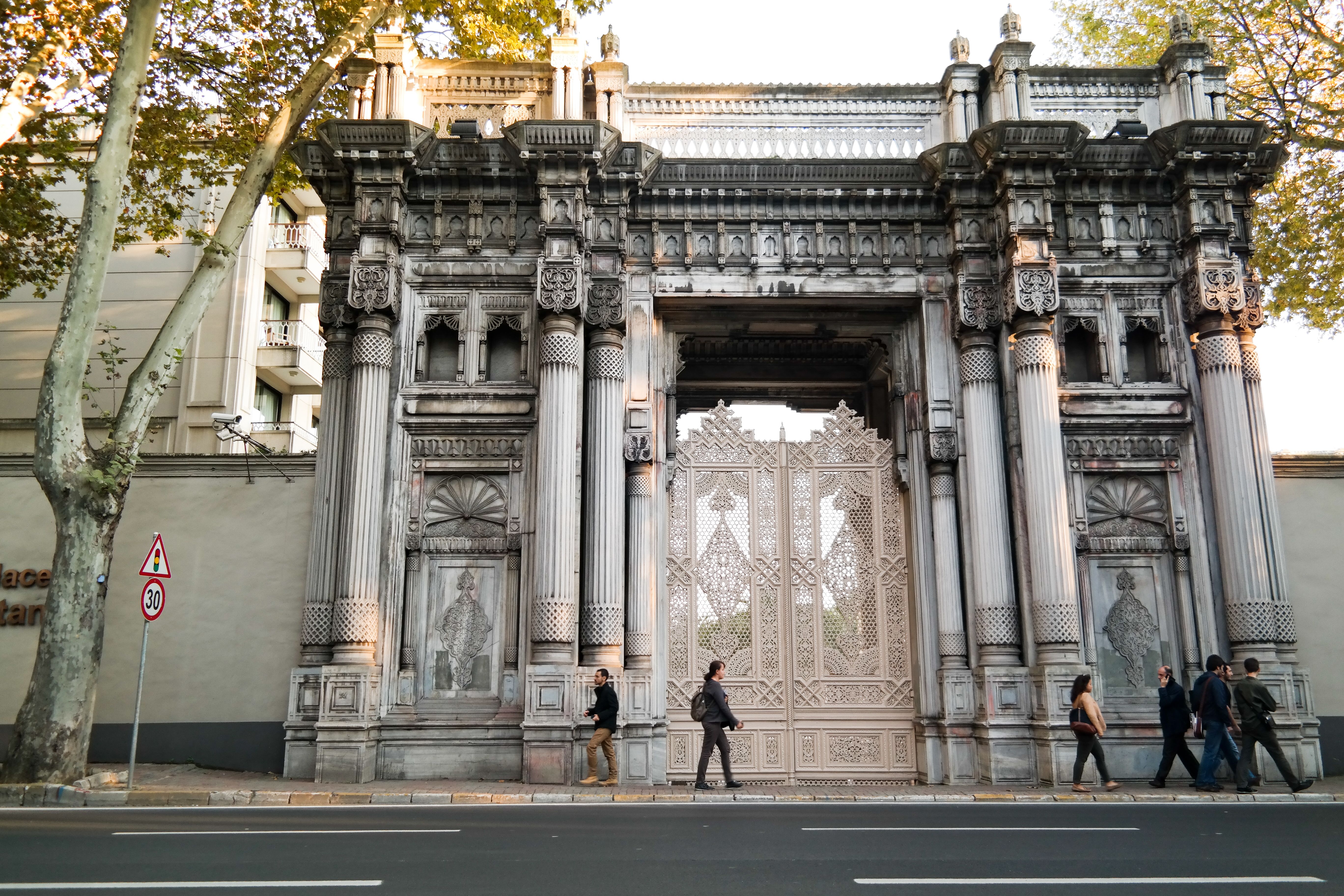 Pedestrians walking by the intricate gate of Ciragan Palace in Istanbul, showcasing baroque architecture.