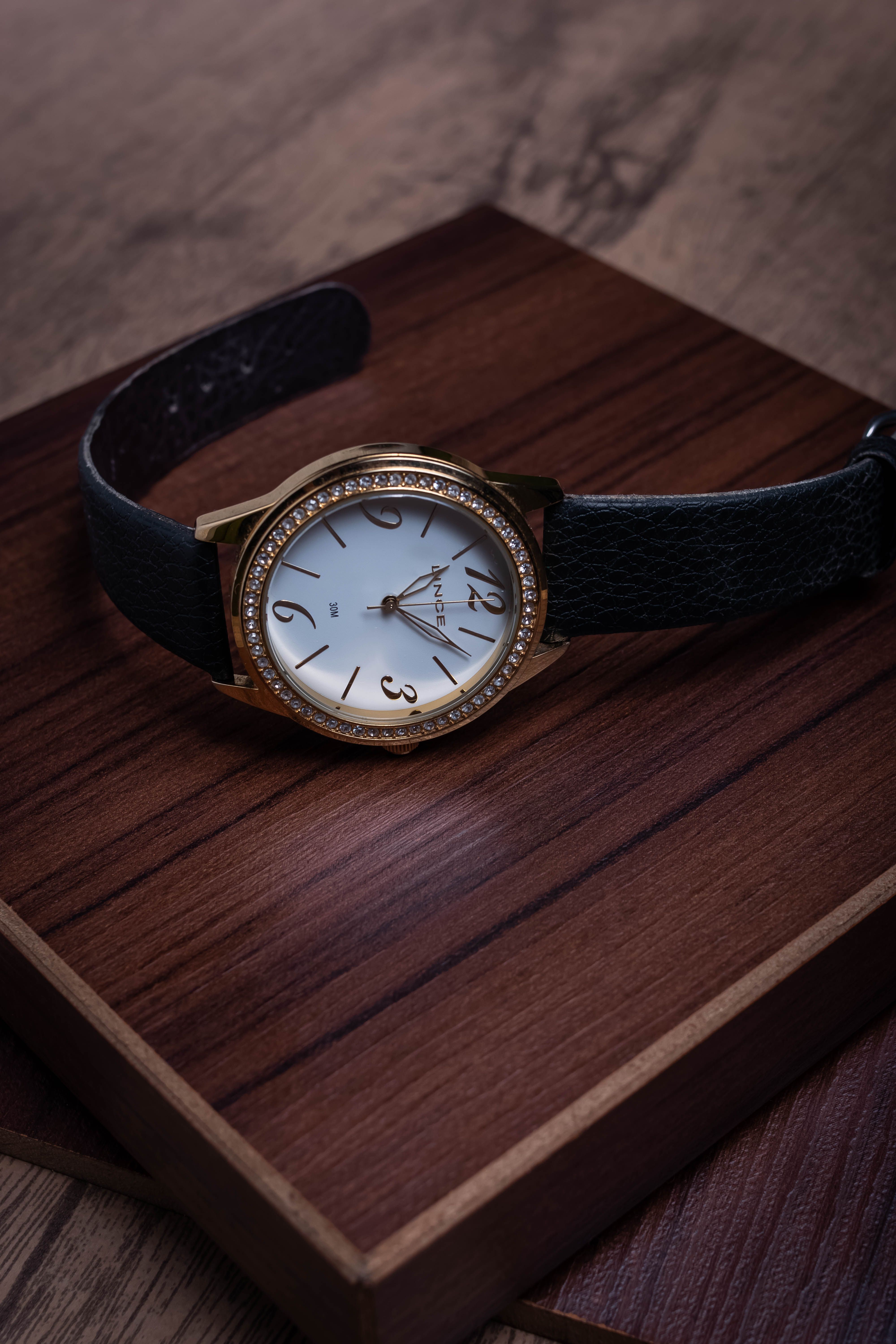 Nox manual watch with gold case and black strap on a wooden tray