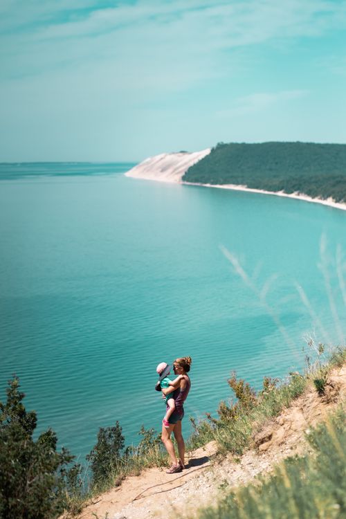Free A mother holds her child overlooking tranquil turquoise waters and scenic dunes on a sunny day. Stock Photo