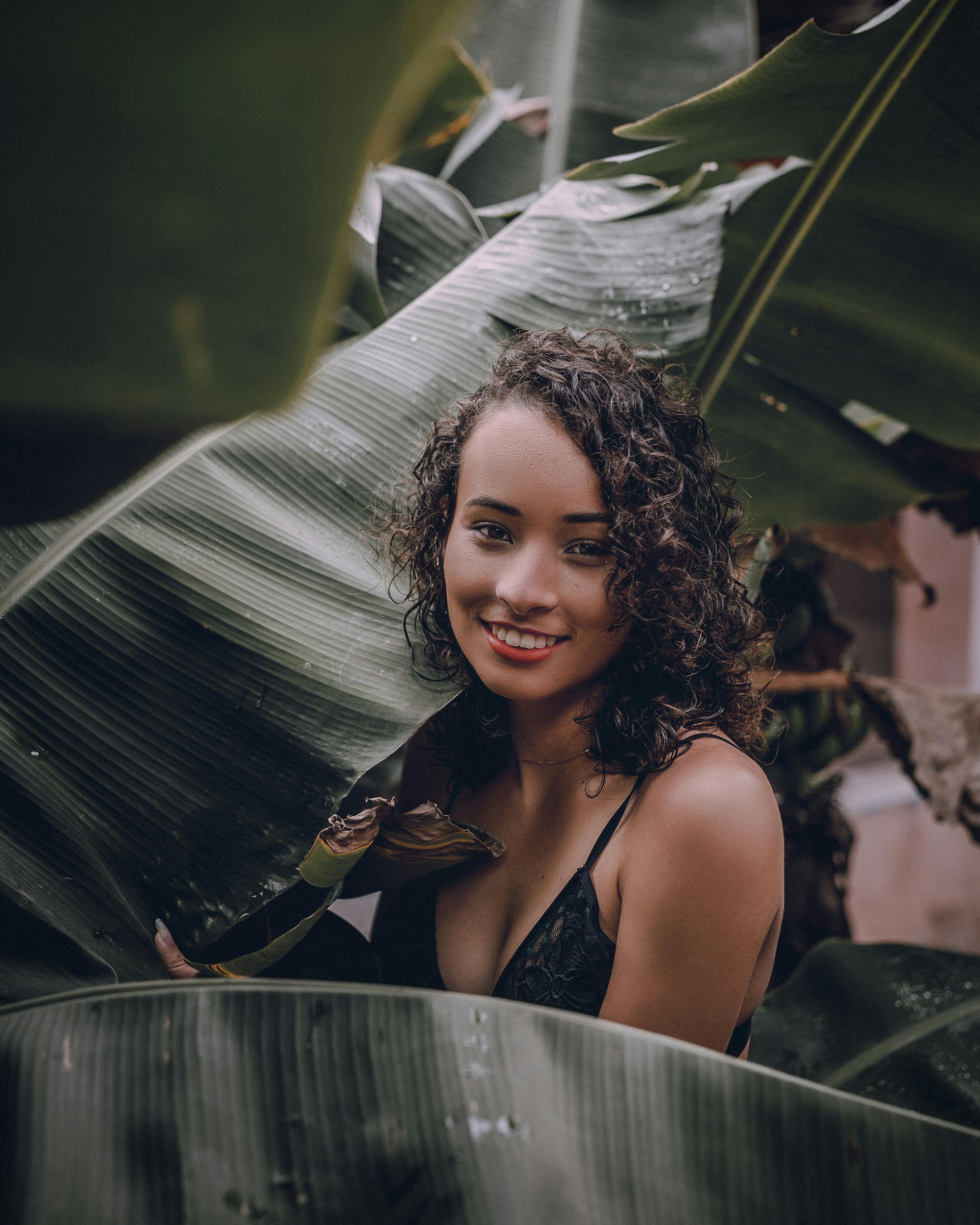 A woman with curly hair joyfully poses among lush tropical leaves in a vertical shot.