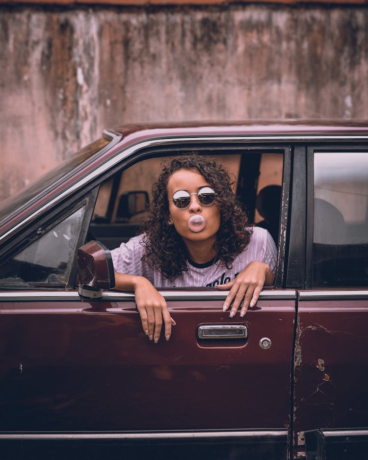 Woman In Sunglasses Blowing A Bubble With Chewing Gum In A Car Window